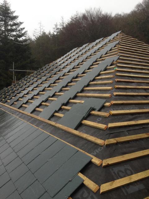 Stacks of dark grey roofing slates distributed across timber battens and black underlay on a hipped roof